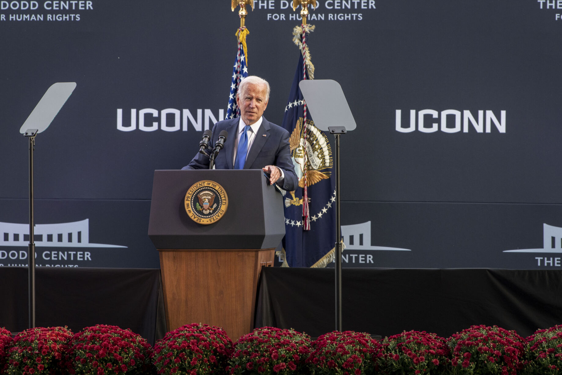 A speaker stands at a presidential podium delivering remarks at a UConn event, with American flags and teleprompters on either side