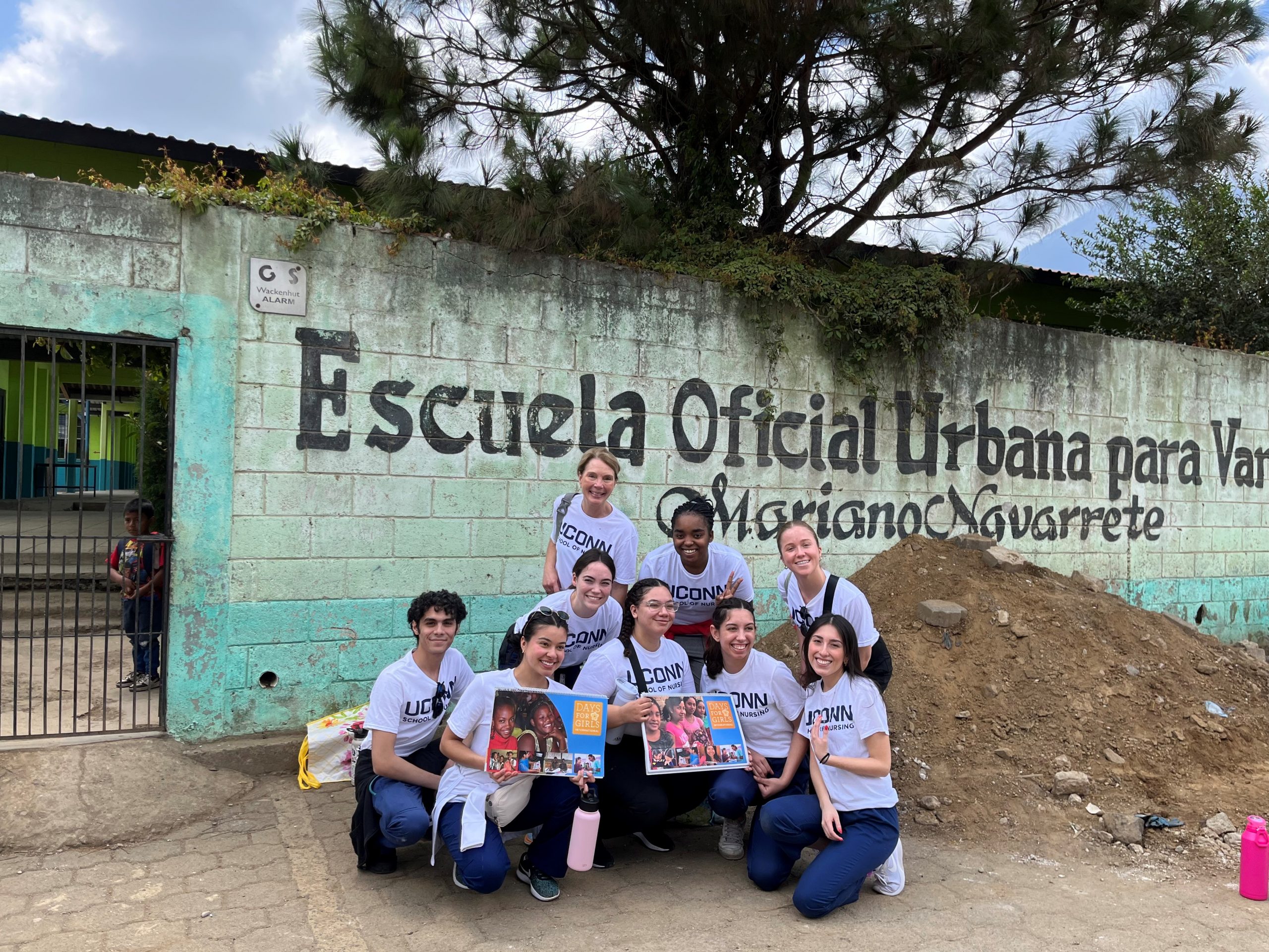 A group of nine people in matching white “UConn School of Nursing” T‑shirts kneel and stand in front of a weathered turquoise and white brick wall with the painted words “Escuela Oficial Urbana para Varones Mariano Navarrete.”