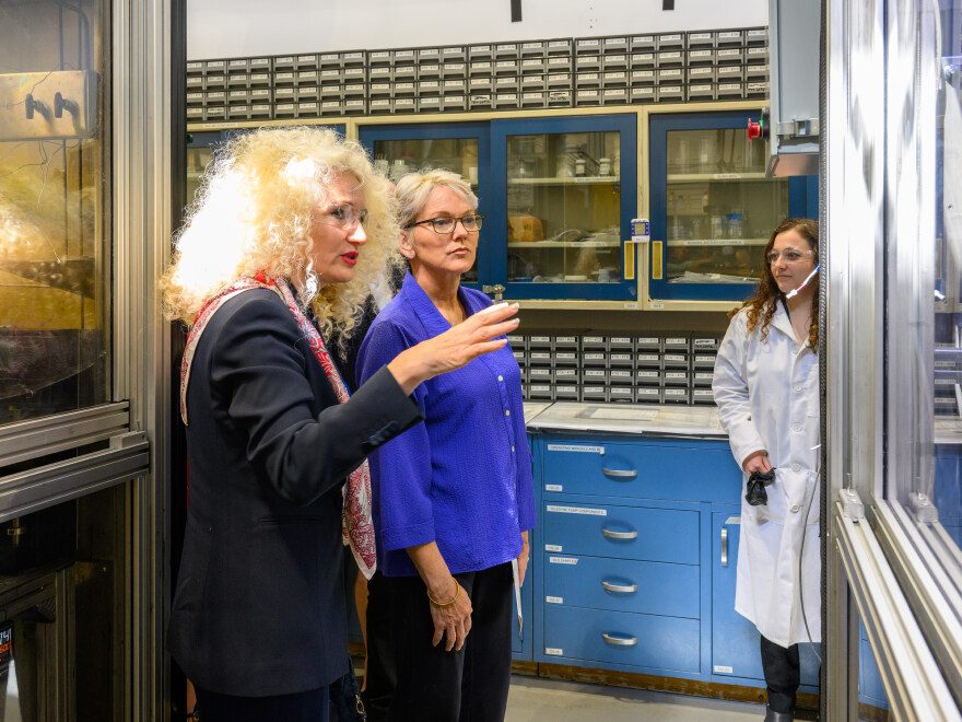 Two women speak during a tour inside a laboratory while a researcher in a lab coat observes nearby.