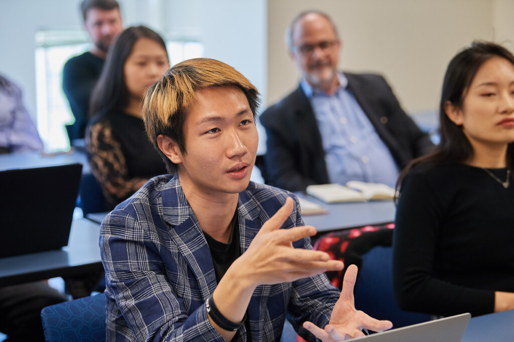 Student speaking and gesturing during a classroom discussion, with other students and a professor listening in the background