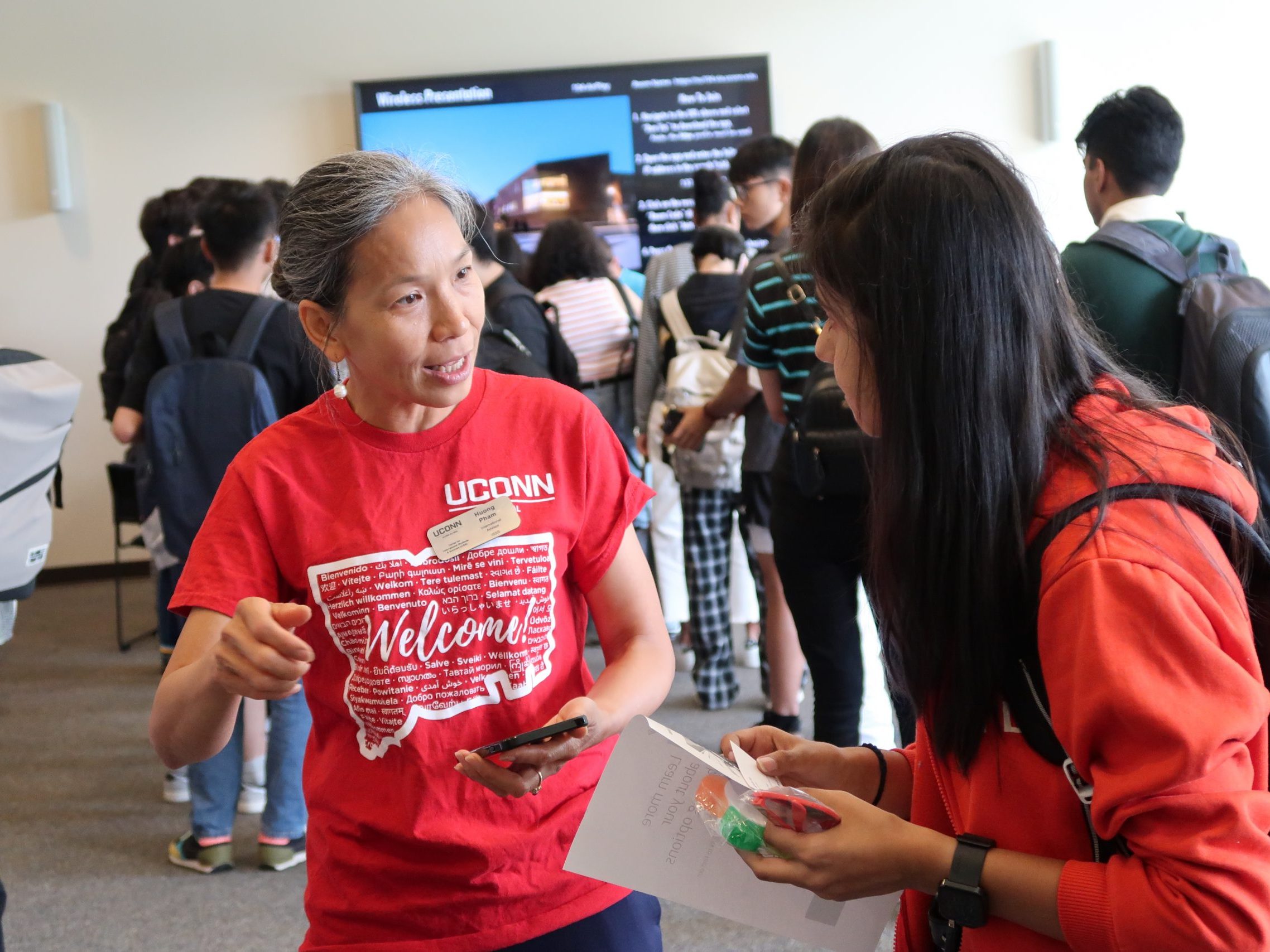 A staff member in a red UConn “Welcome” T‑shirt speaks with a student in an orange hoodie holding papers at a busy campus orientation or reception, with other students and an information screen in the background.
